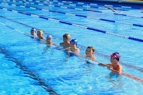 A photo of a group of children in a pool resting on the lane divider and smiling at the camera.